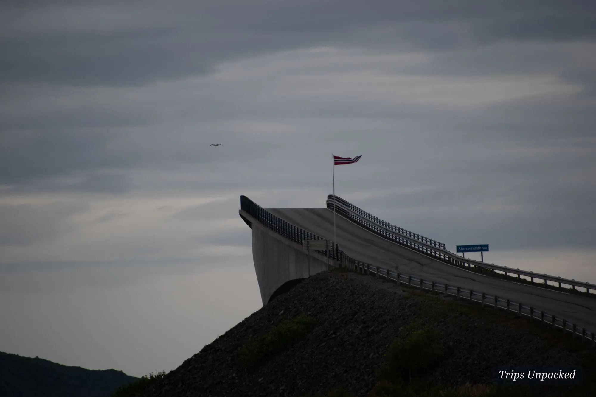 The Storseisund bridge of the Atlantic Ocean Road curving into a grey sky, a Norwegian flag flying.