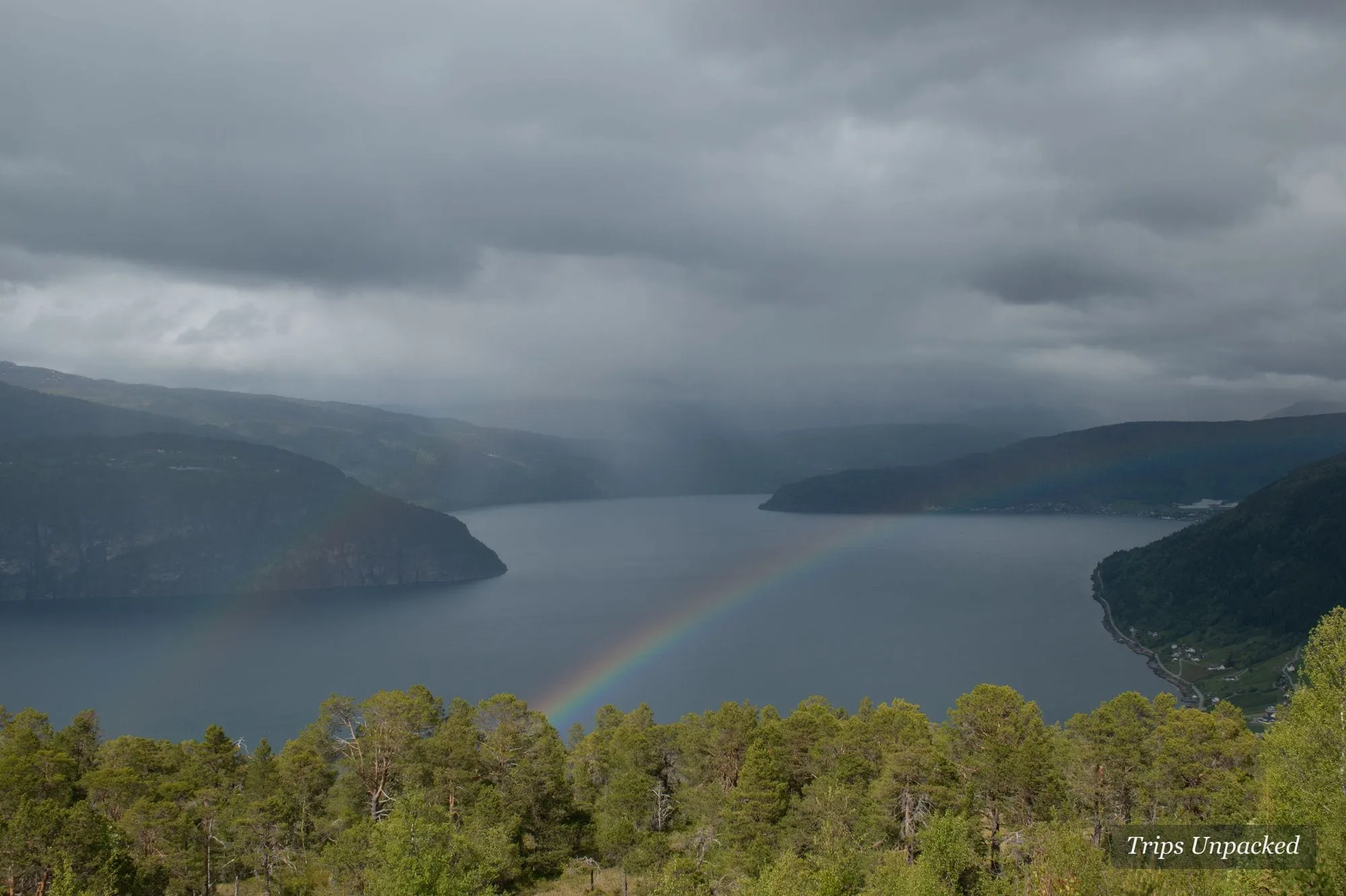 A rainbow arcing over a broad fjord under a rain-heavy sky.