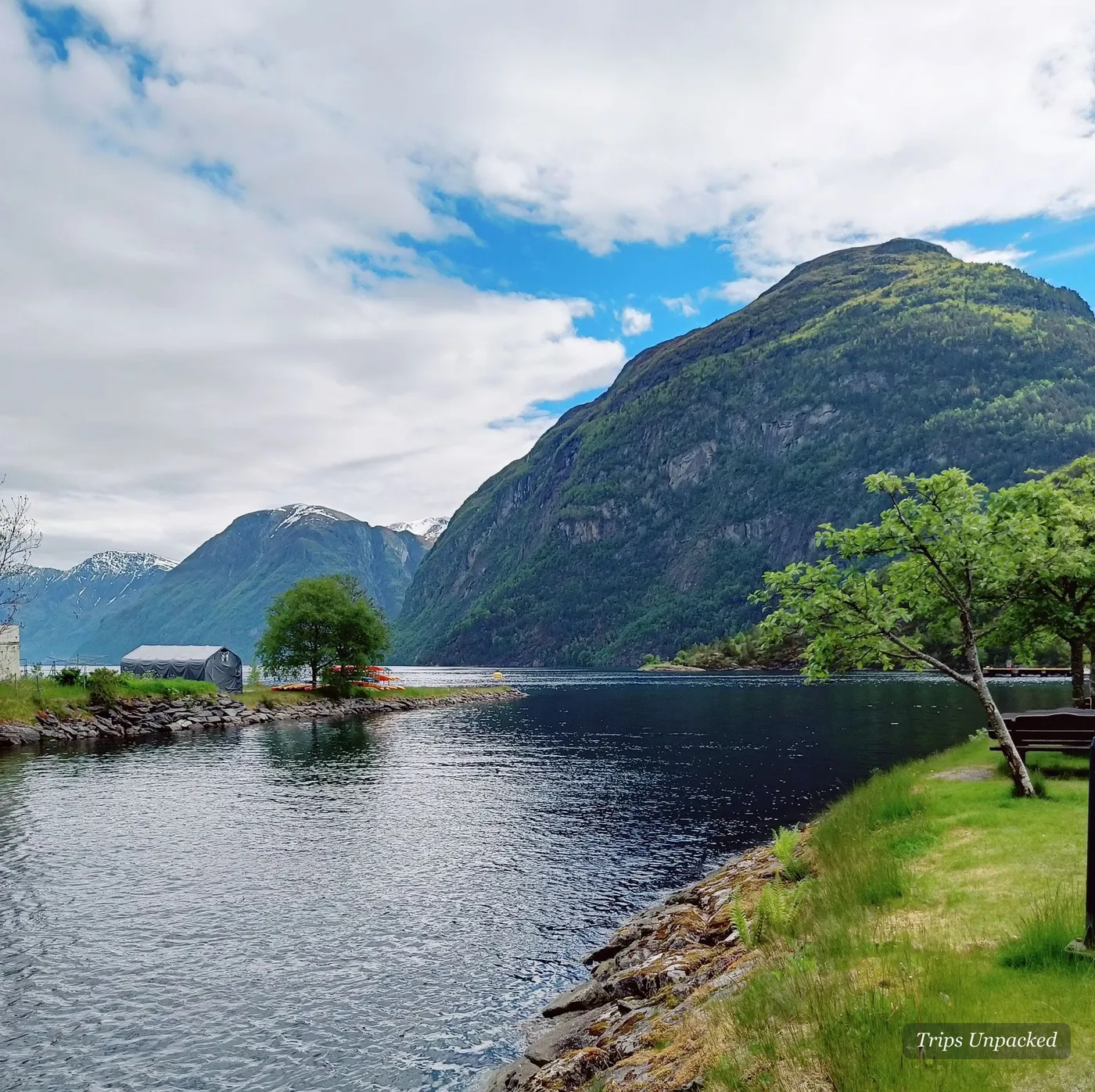 A quiet fjord inlet with kayaks, a lone tree and a bench on the grassy shore.