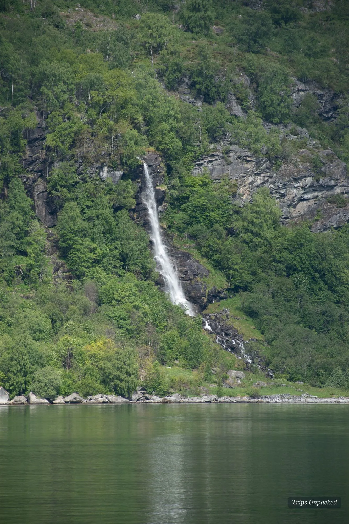 A slender waterfall threading down a forested cliff into the Geirangerfjord.